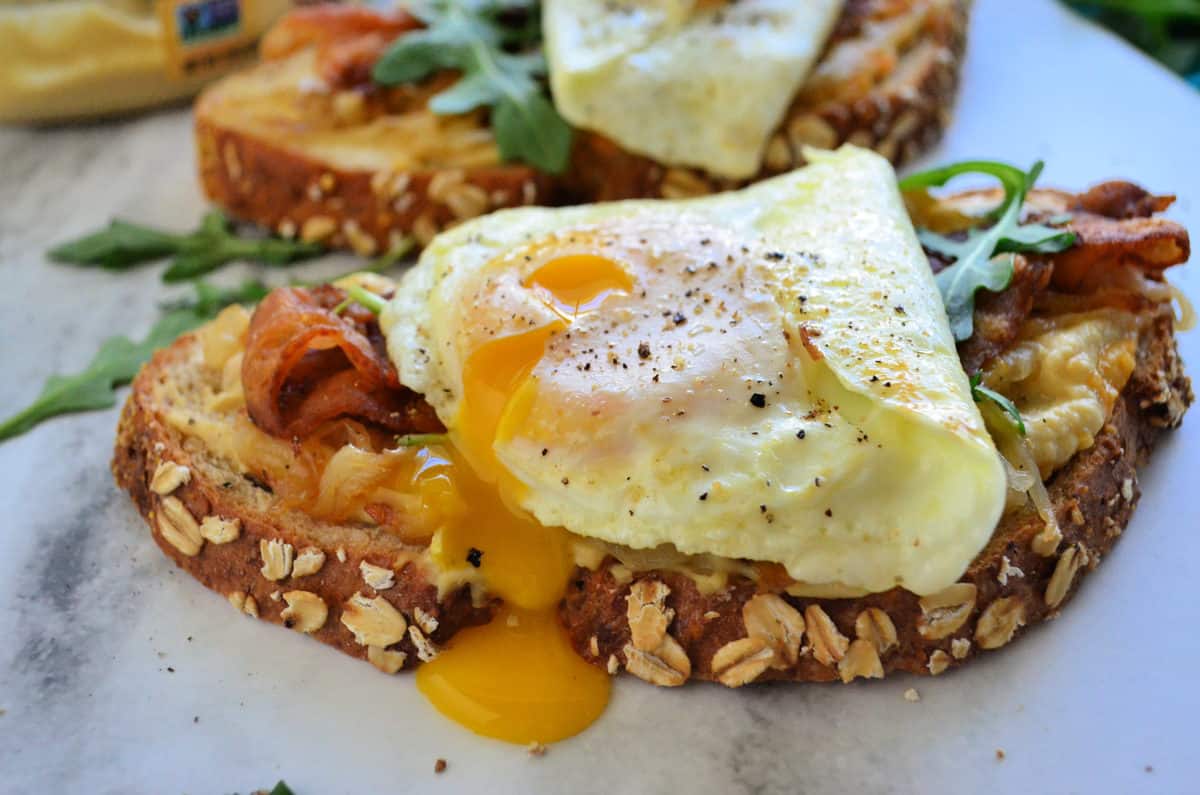 bread topped with hummus, bacon, over easy eggs oozing yolk, and arugula on platter.