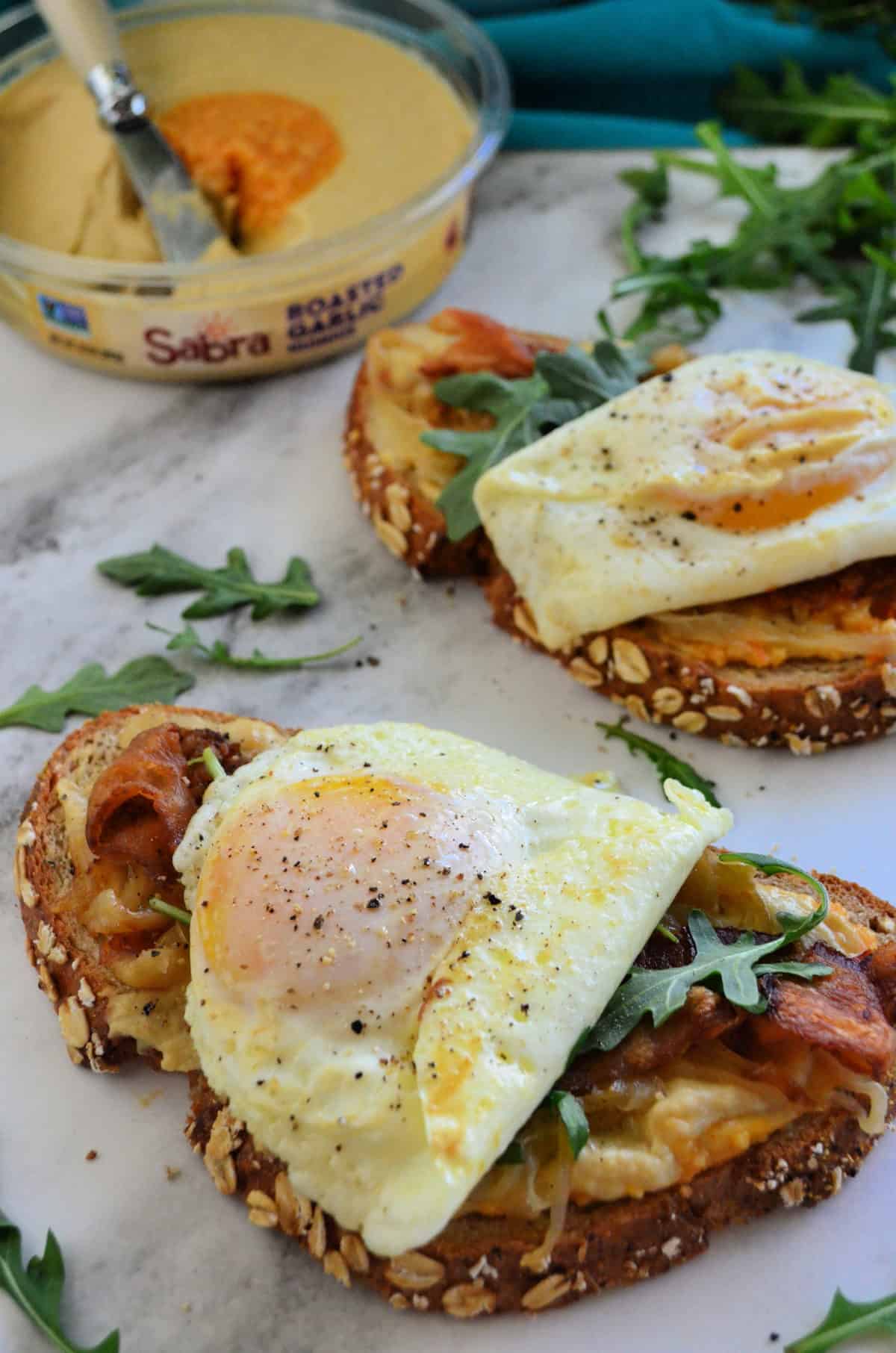 bread with hummus, bacon, over easy eggs oozing yolk, and arugula on platter with hummus behind.
