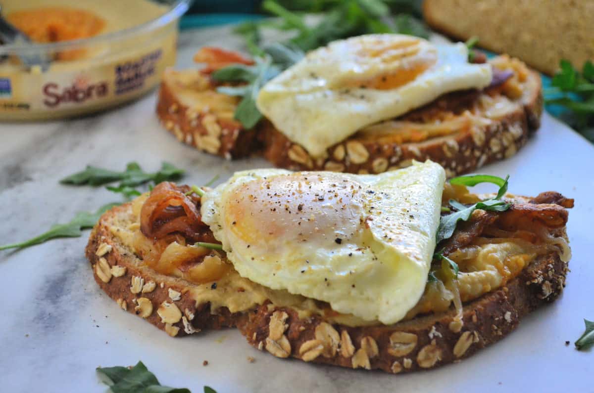 side view hummus container, bread topped with hummus, bacon, over easy eggs, arugula on platter.