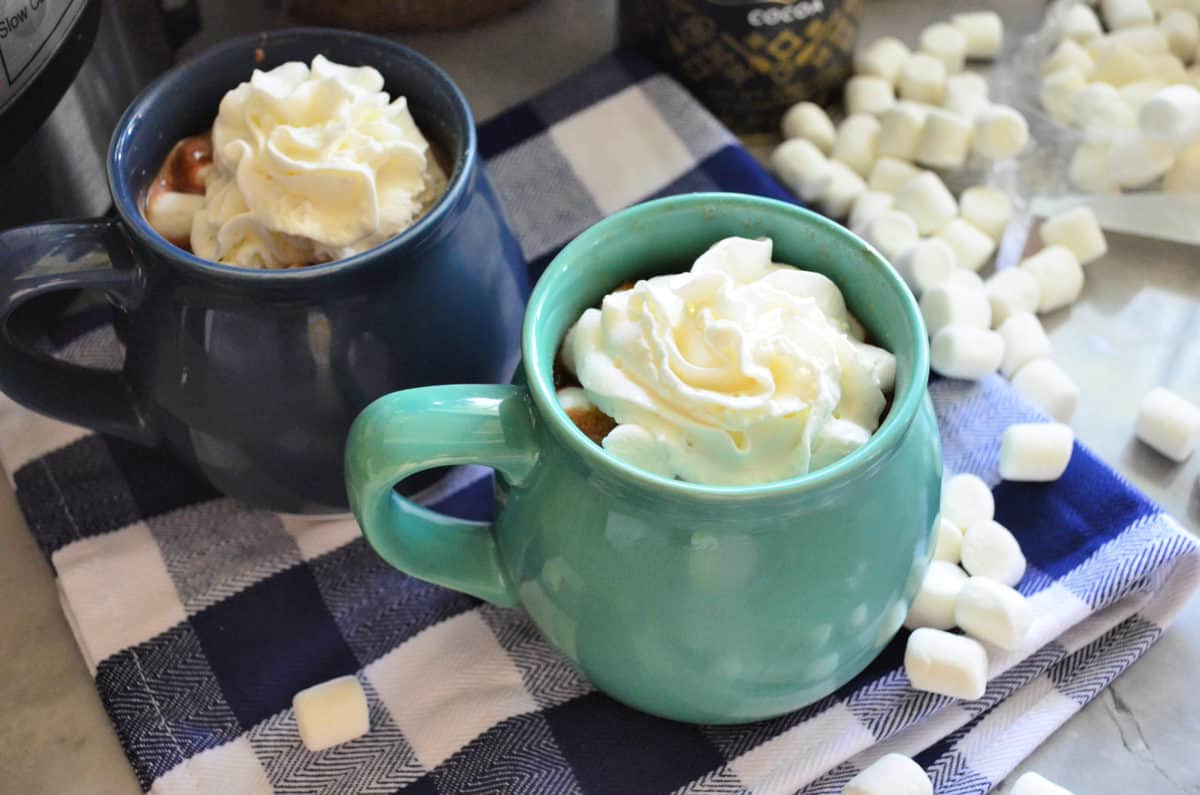 two mugs with whipped cream showing from the top on checkered tablecloth with mini marshmallows.