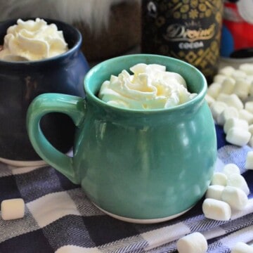 two mugs on tablecloth with whipped cream showing from the top of the mugs.