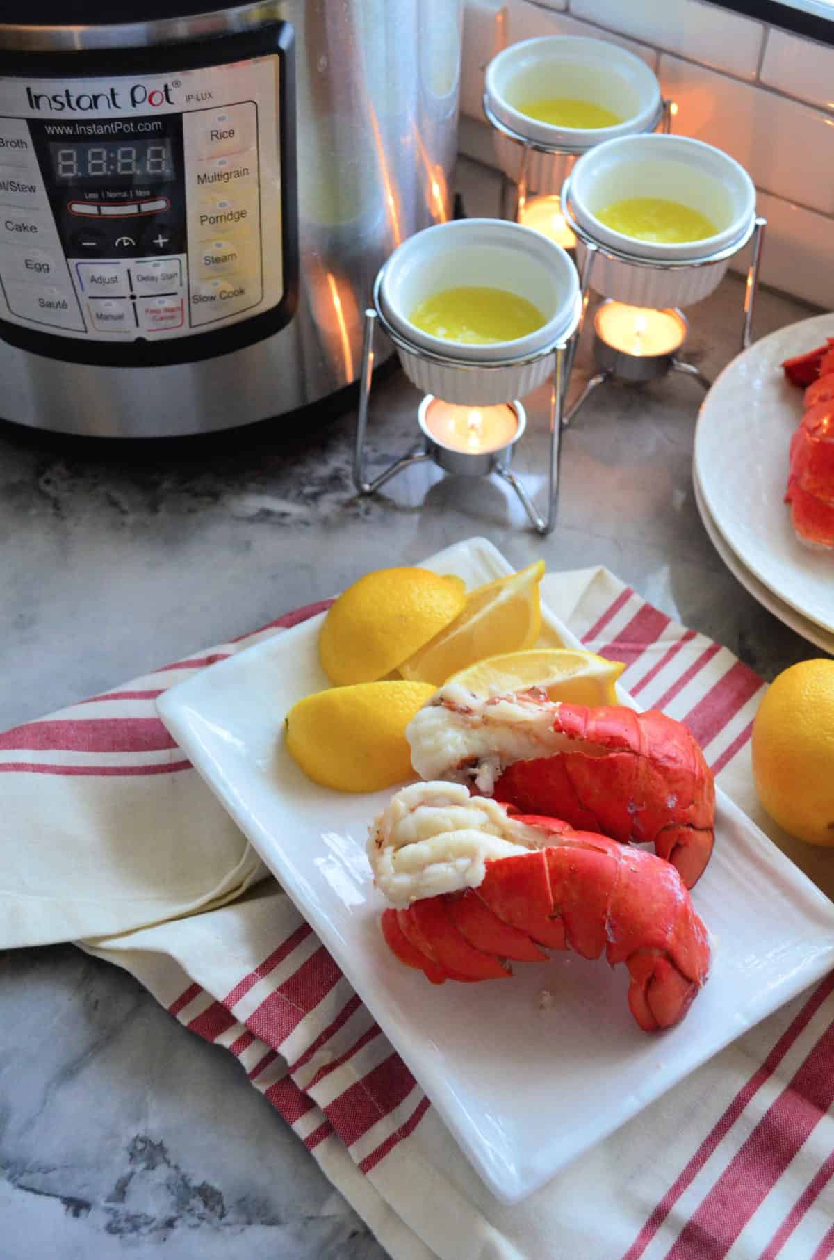 Two plated lobster tails with lemon and hot butter on tablecloth in front of instant pot.