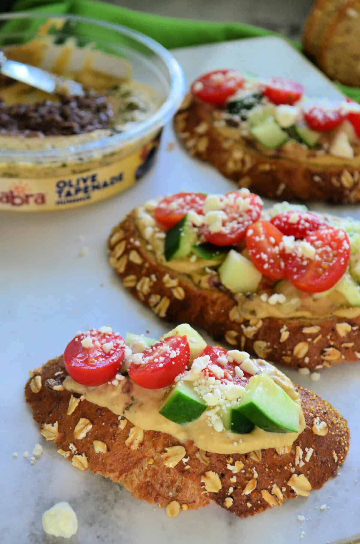 3 toast slices topped with hummus, cucumber, tomato, and feta with sabra hummus blurred behind.