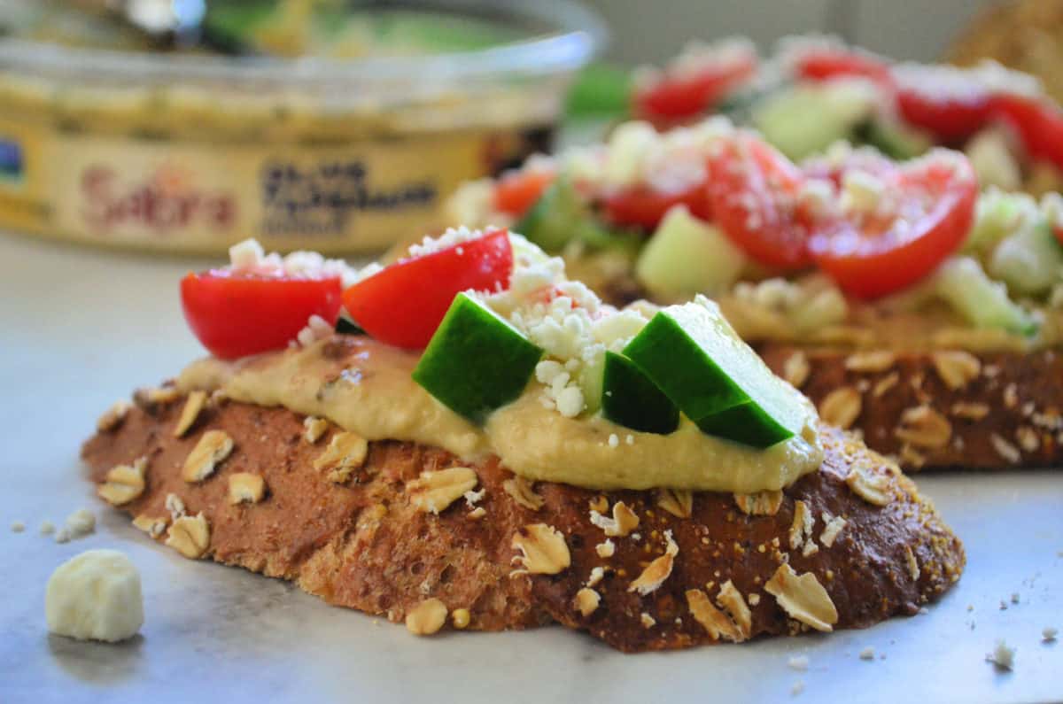 closeup side view of toast slice topped with hummus, cucumber, tomato, and feta.