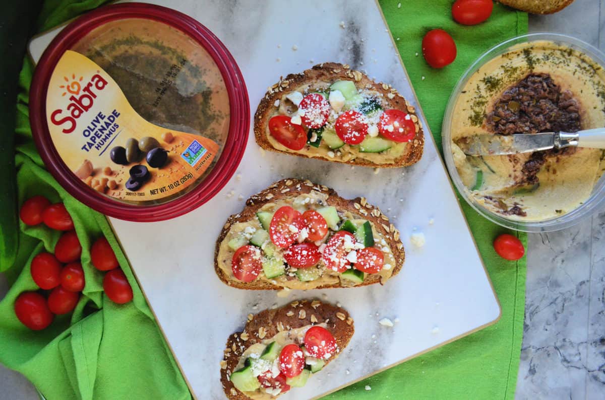 3 toast slices topped with hummus, cucumber, tomato, and feta next to Sabra hummus container.