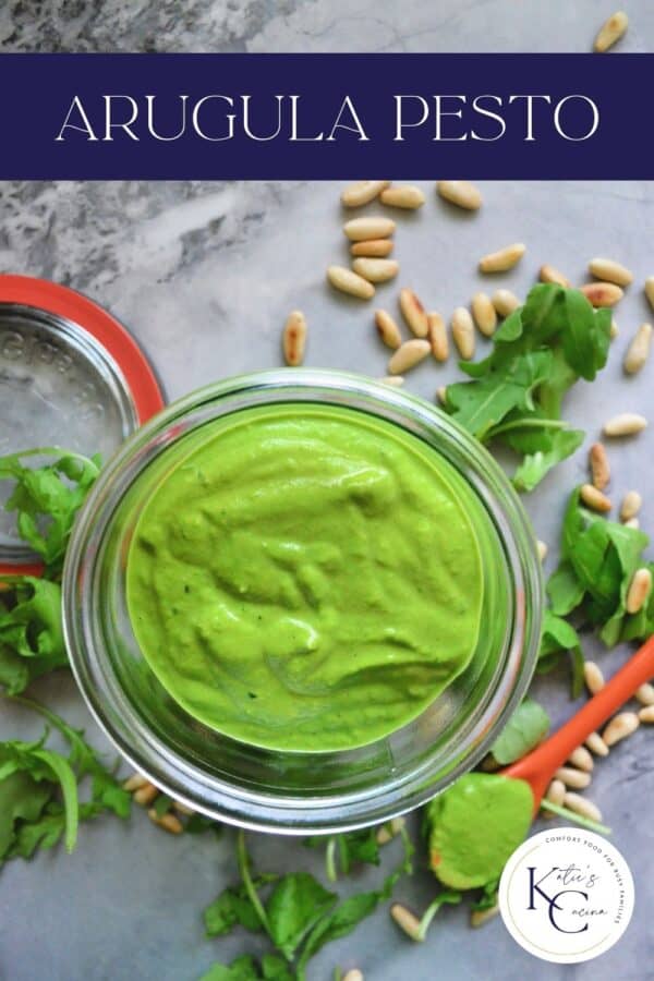 Top view of a glass jar with a green sauce and arugul and pine nuts on counter with text on image for Pinterst.