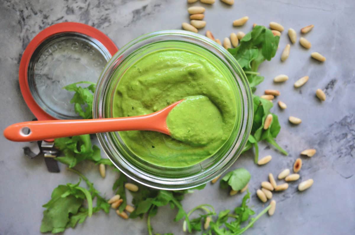 top view glass bowl with thick green sauce on counter with arugula and pine nuts. 