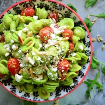 Top view of bowl of pasta in green sauce with tomatoes, cheese crumbles, and pine nuts on counter with arugula.