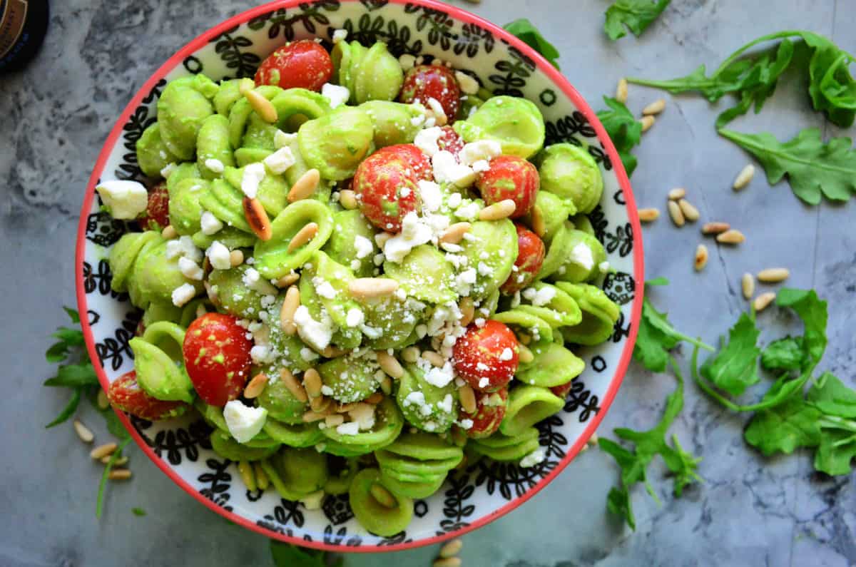 top view bowl of orrichette pasta in green sauce with pine nuts, feta, grape tomatoes on counter.