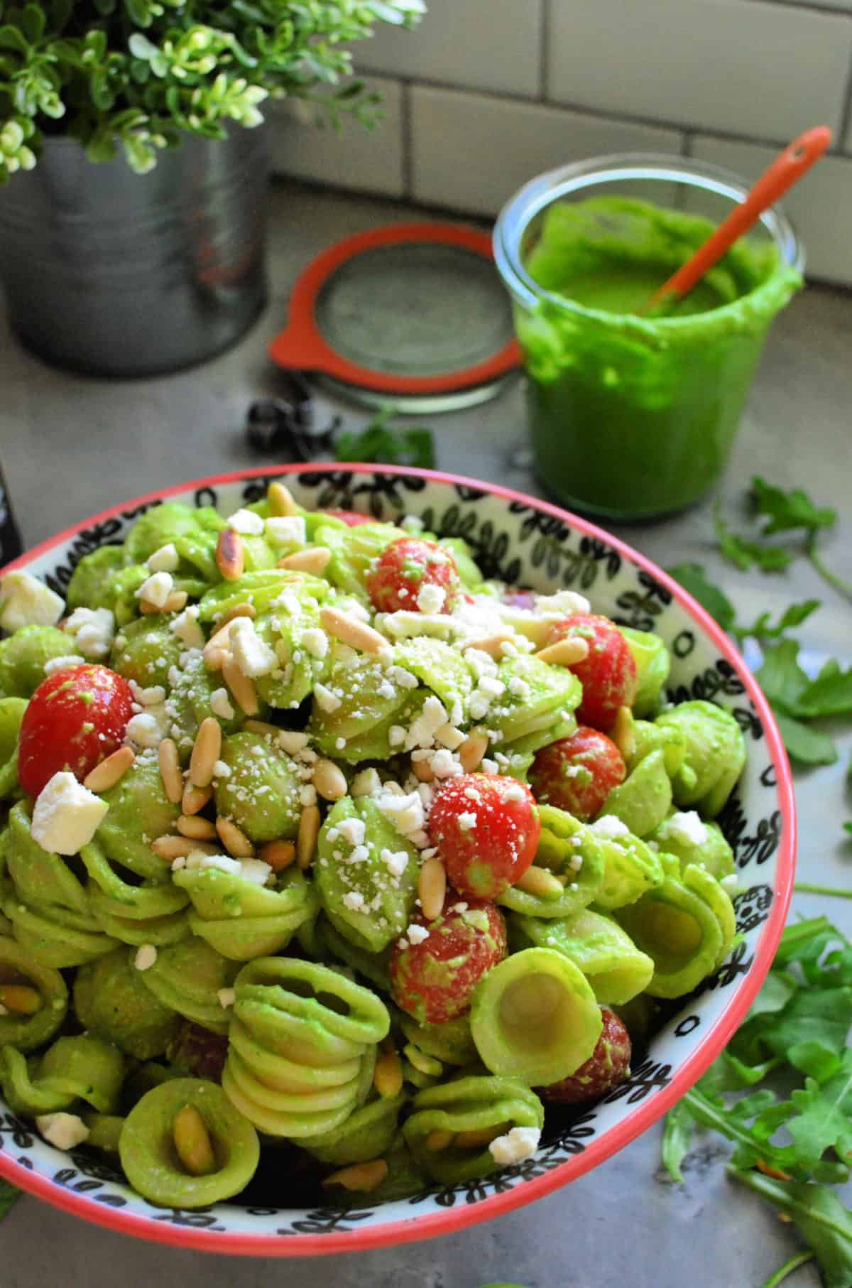 Bowl of pasta shells in green sauce topped with tomato, pine nuts, cheese crumbles on countertop.