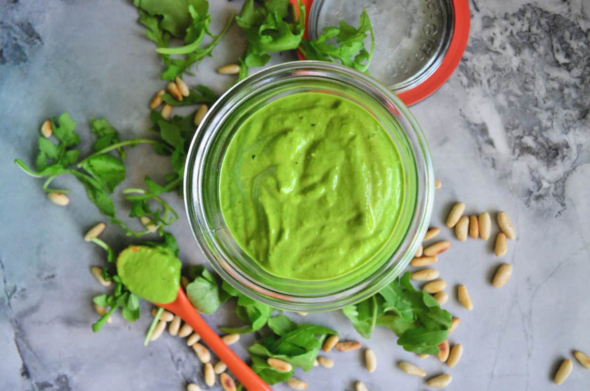 top view glass bowl with thick green sauce on counter with arugula, pine nuts, and spoon.