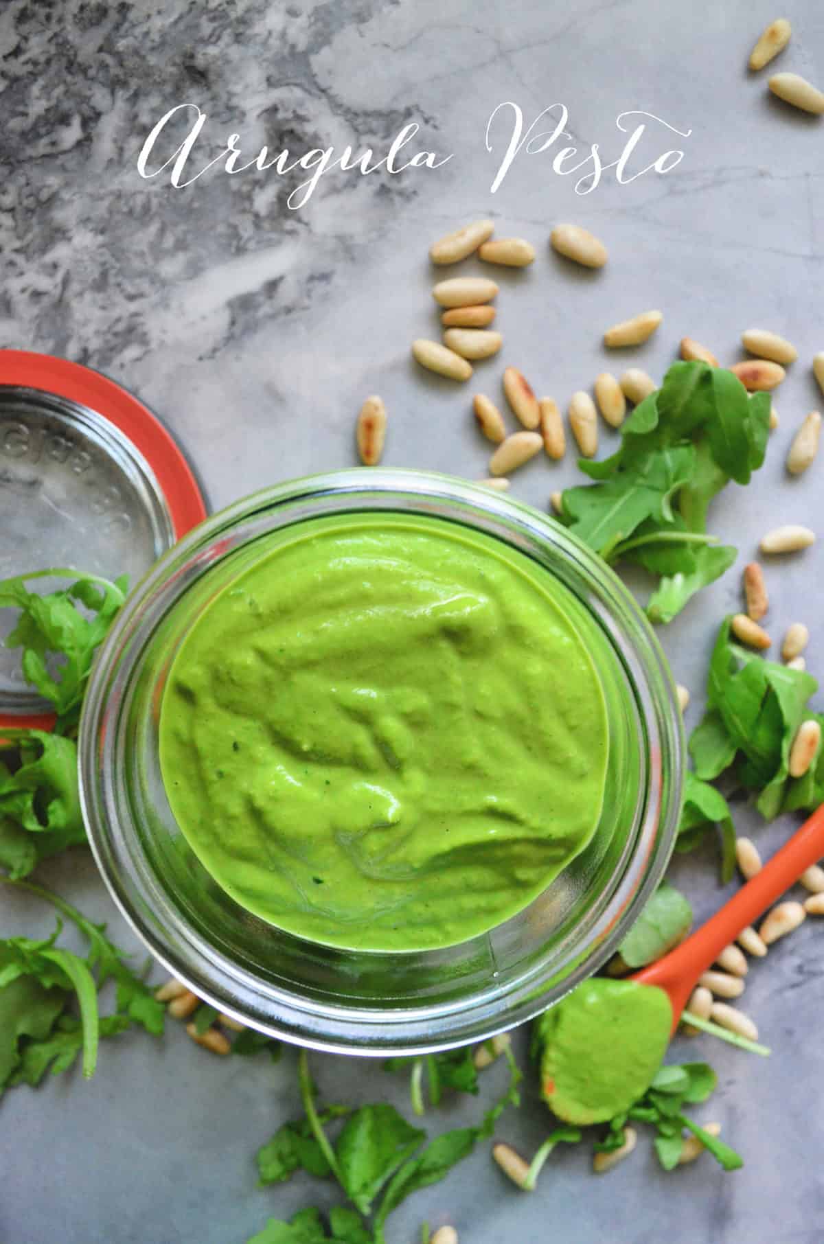 top view glass bowl with thick green sauce on counter with arugula and pine nuts. Title Text.