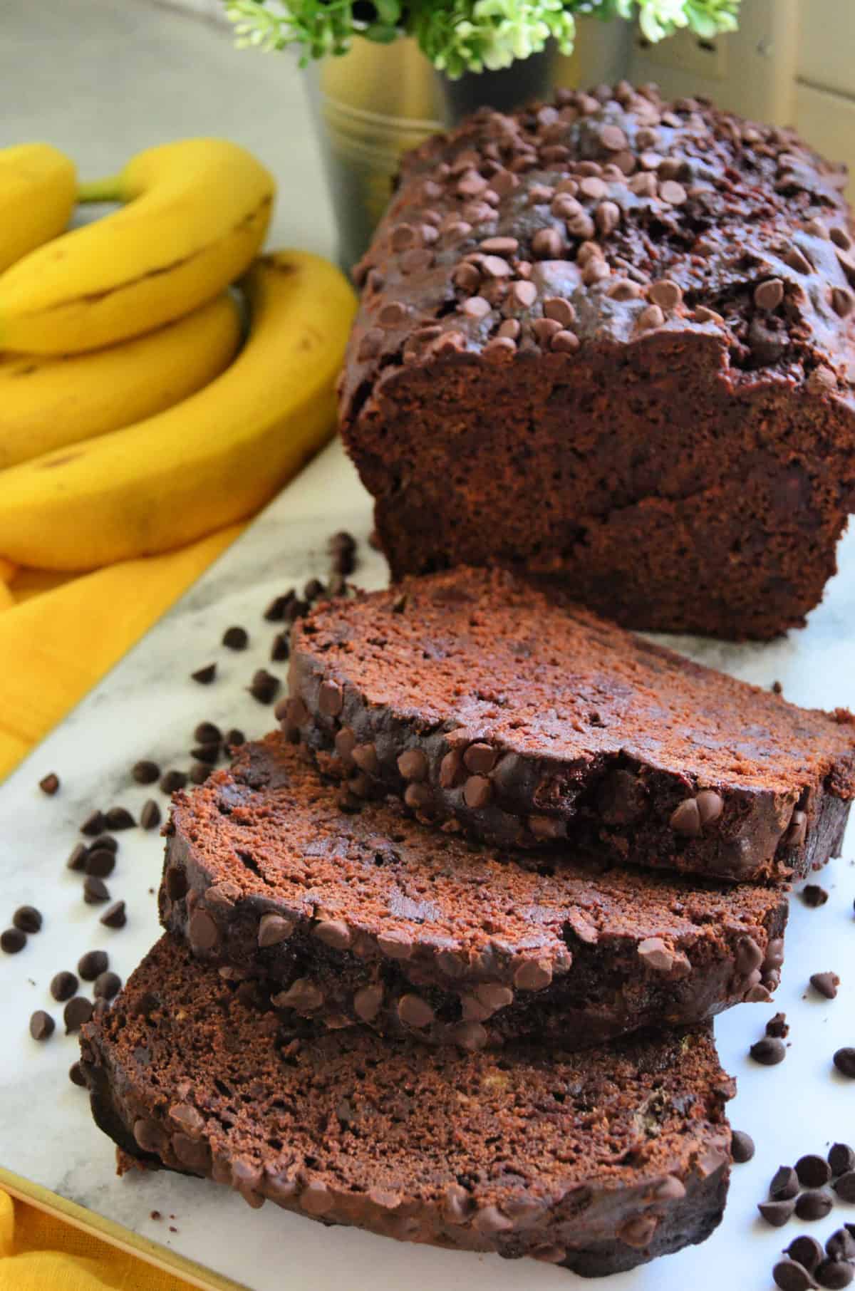 3 slices of chocolate chocolate chip bread and loaf on countertop with bananas in background.