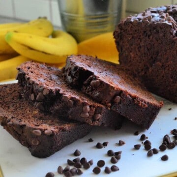 loaf of chocolate chocolate chip banana bread on yellow tablecloth with 3 slices laid out.