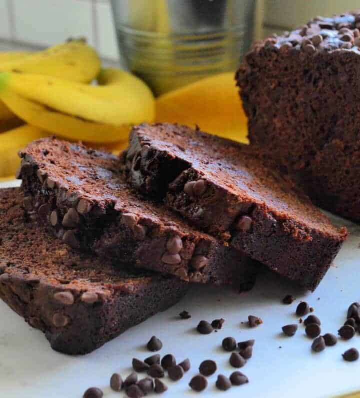 loaf of chocolate chocolate chip banana bread on yellow tablecloth with 3 slices laid out.