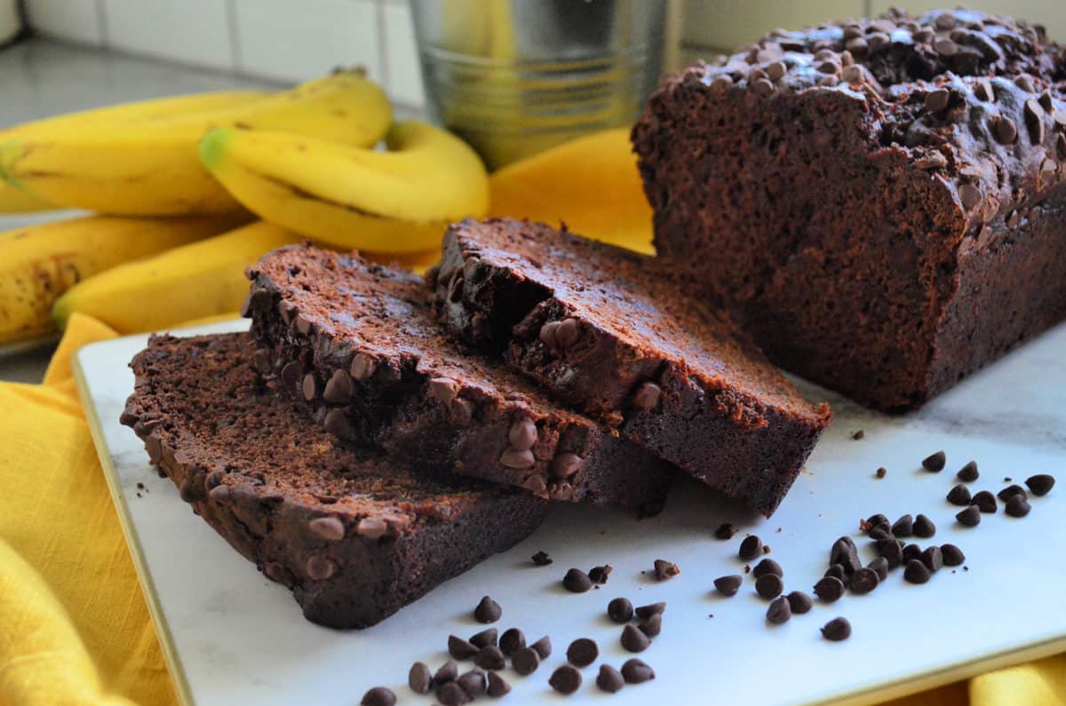 3 slices of chocolate chocolate chip bread on countertop in front of bananas with remaining loaf behind.