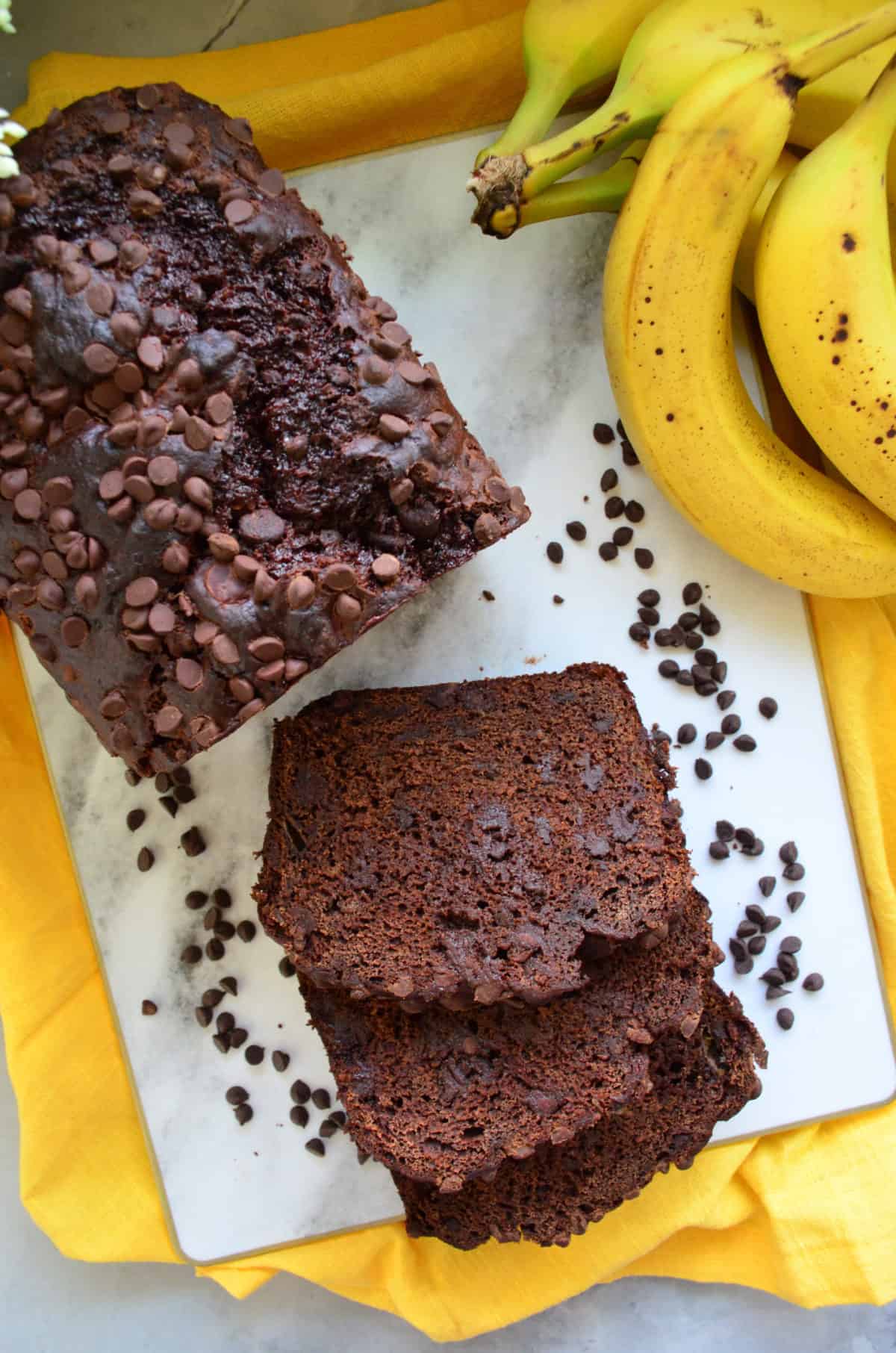 3 slices of chocolate chocolate chip bread and loaf on countertop with chocolate chips on yellow cloth.