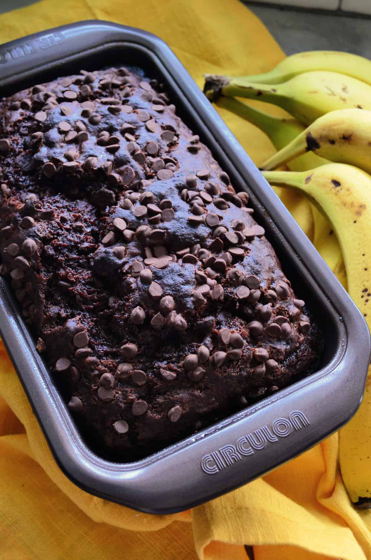 Top view of loaf of brown bread in pan with chocolate chips in it and bananas in background.