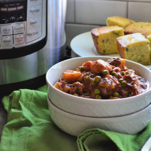 bowl of thick beef stew with potatoes, carrots, celery, and peas visible in front of plated cornbread.