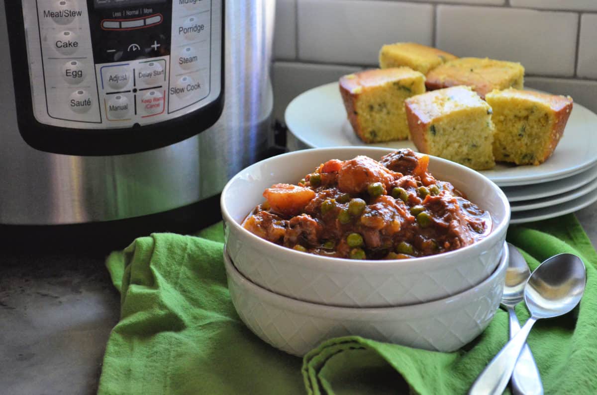 bowl of thick beef stew with potatoes, carrots, celery, and peas visible in front of plated cornbread.