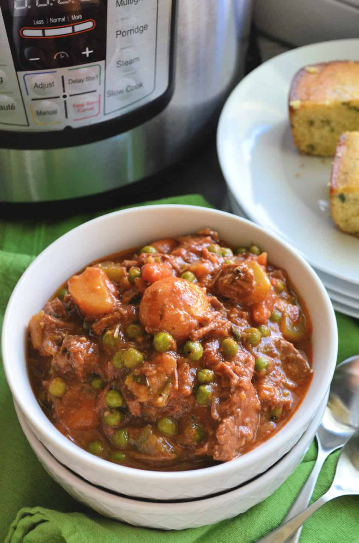Bowl of beef stew with visible potatoes, peas, and carrots in front of instant pot and corn bread.