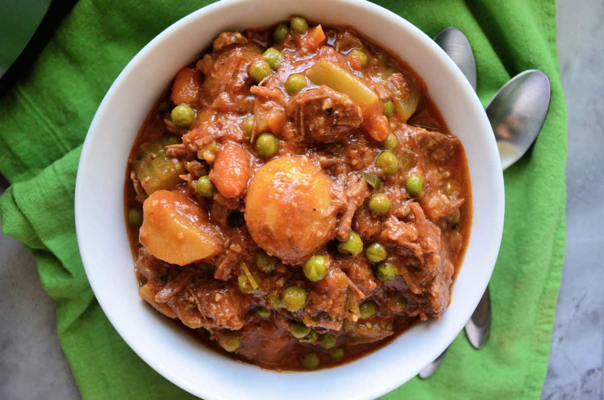 top view bowl beef stew with visible potatoes, peas, and carrots with corn bread behind it.