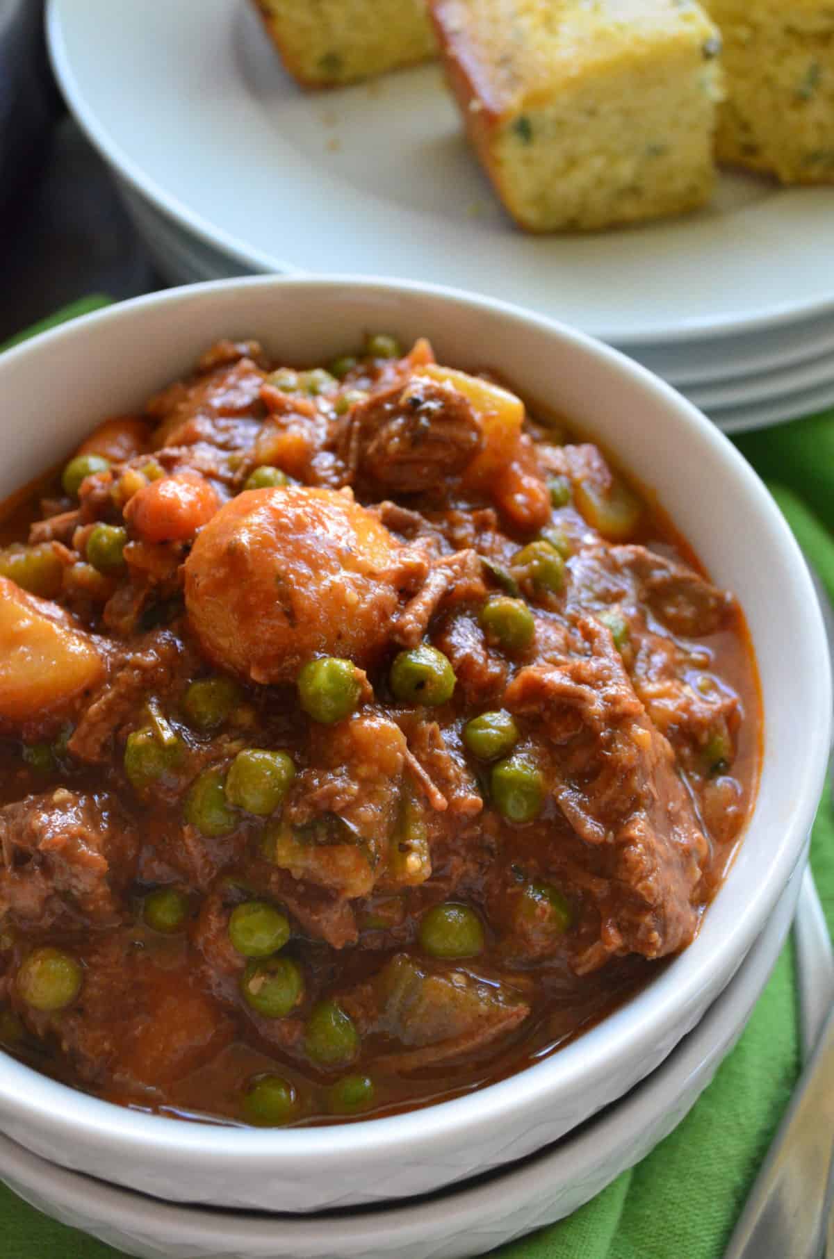 Closeup bowl beef stew with visible potatoes, peas, and carrots with corn bread behind it.