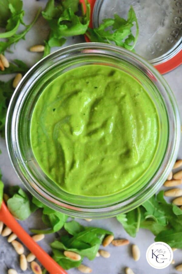 Close up of a glass jar with bright green sauce and pine nuts on counter.