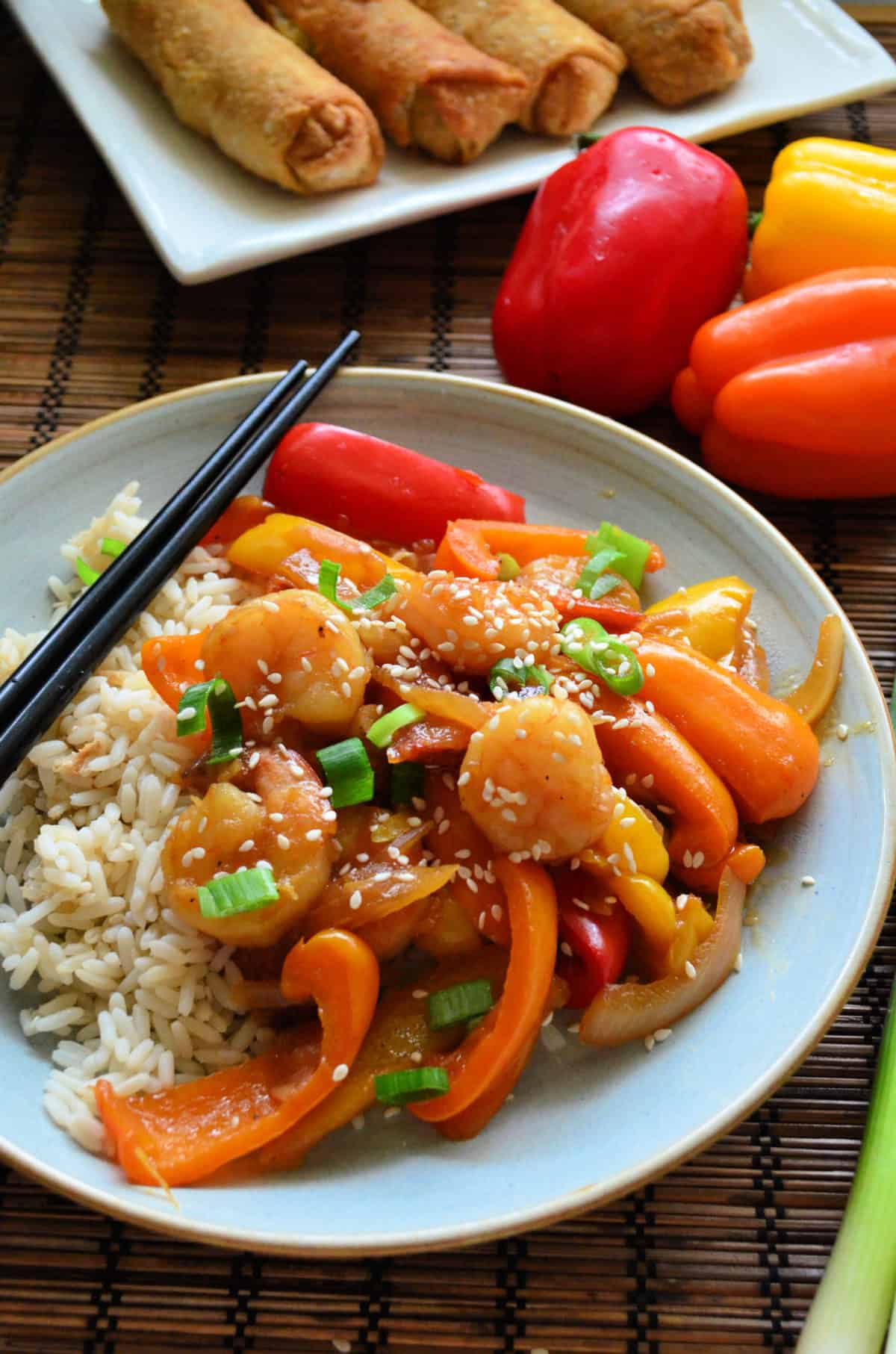 Plated sauce, shrimp, peppers, green onions, and sesame seeds over plated rice bed near peppers.