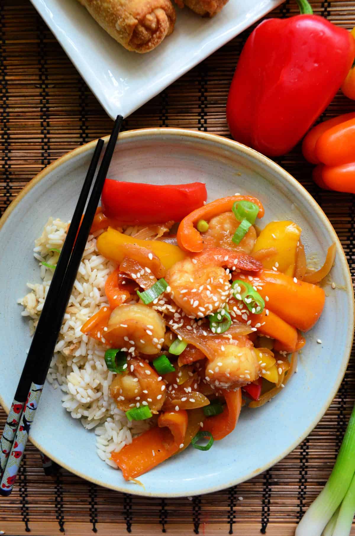 Plated sauce, shrimp, peppers, green onions, and sesame seeds over plated rice bed with chopsticks.