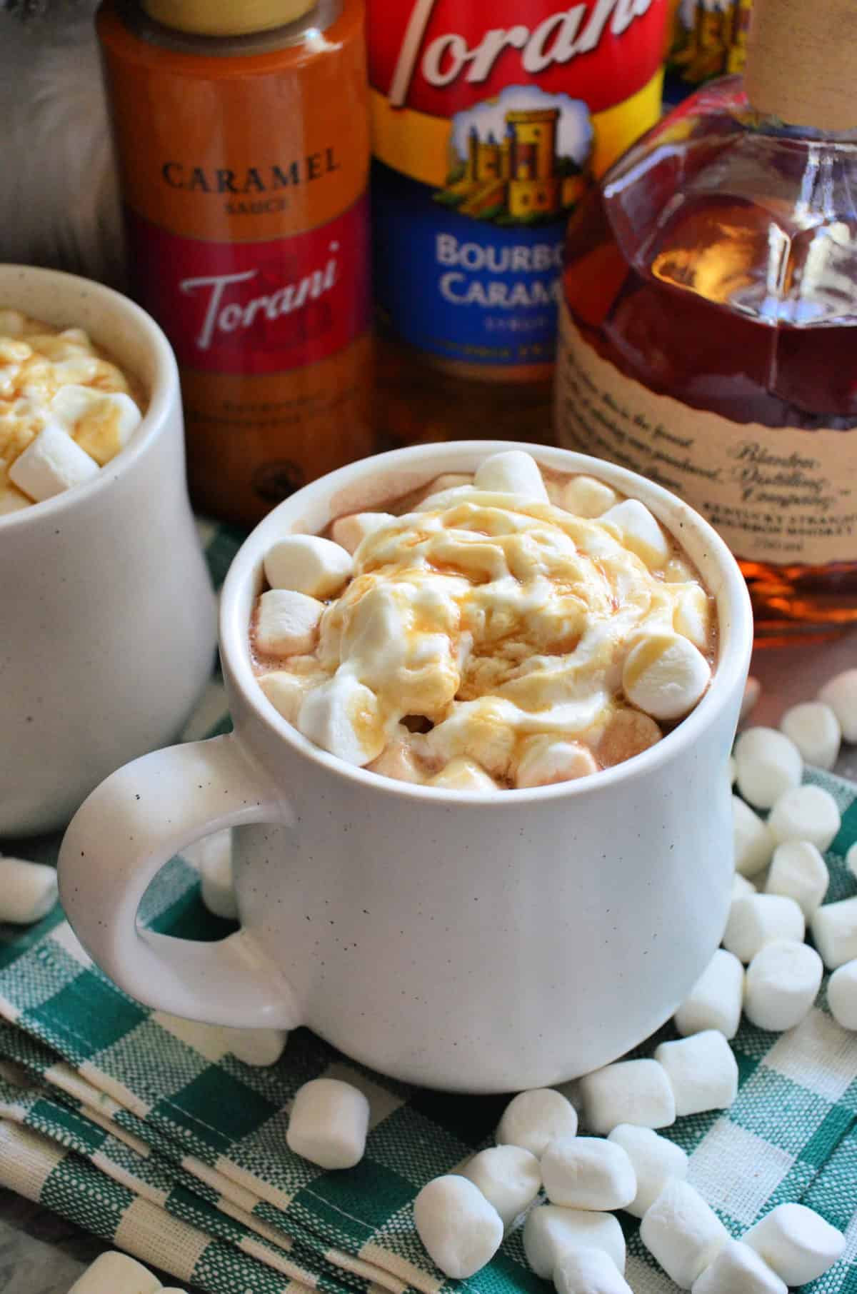 two mugs of brown liquid topped with mini marshmallows, whipped cream, and caramel with ingredients behind.