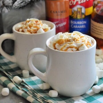 Two mugs on tablecloth with whipped cream and caramel drizzle showing from mug tops.