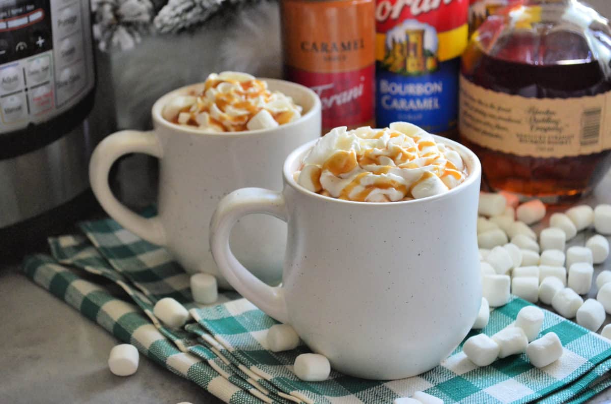 two mugs of brown liquid topped with mini marshmallows, whipped cream, and caramel with ingredients behind.