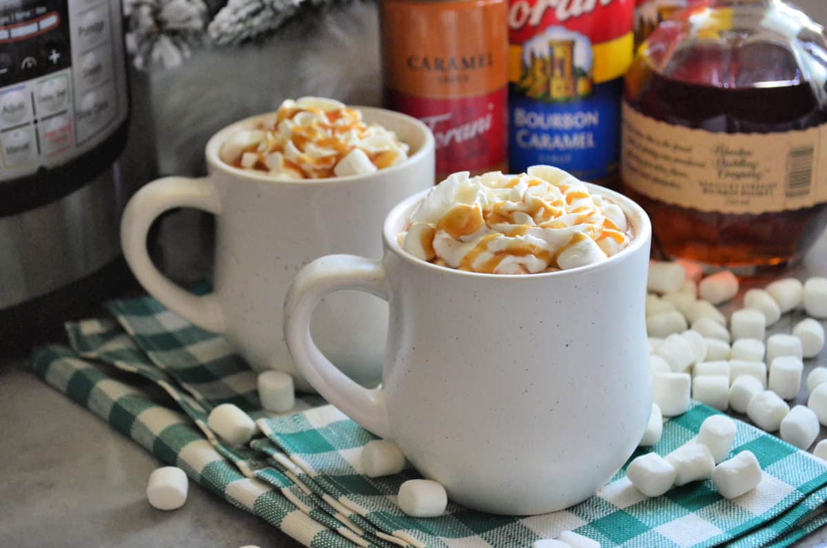 Two mugs on tablecloth with whipped cream and caramel drizzle showing from mug tops.