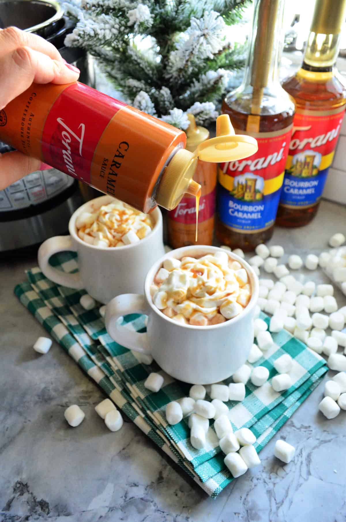 Hand drizzling caramel sauce over mug of liquid with whipped cream and mini mushrooms.