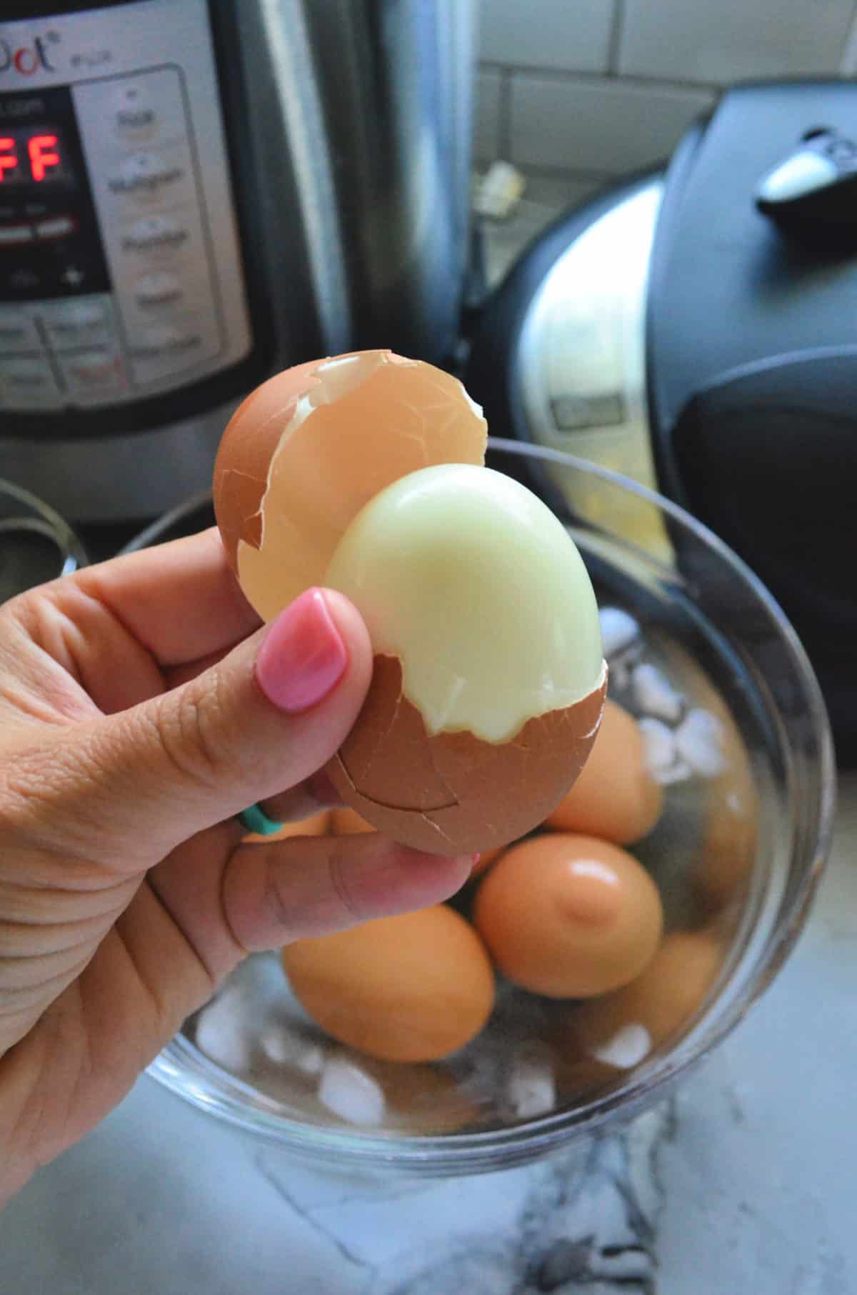Hand holding up hard boiled egg with top portion of shell removed in front of instant pot.