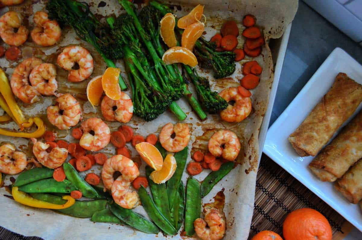 peas, broccolini, mandarine oranges, shrimp, bell pepper, and carrots on paper on baking sheet.