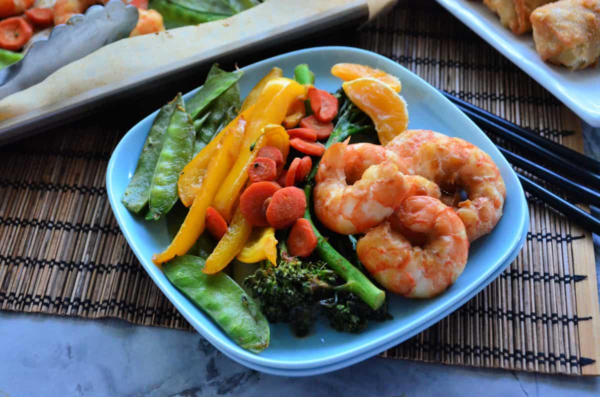 peas, broccolini, mandrine oranges, shrimp, bell pepper, and carrots on paper on plate with chopsticks.