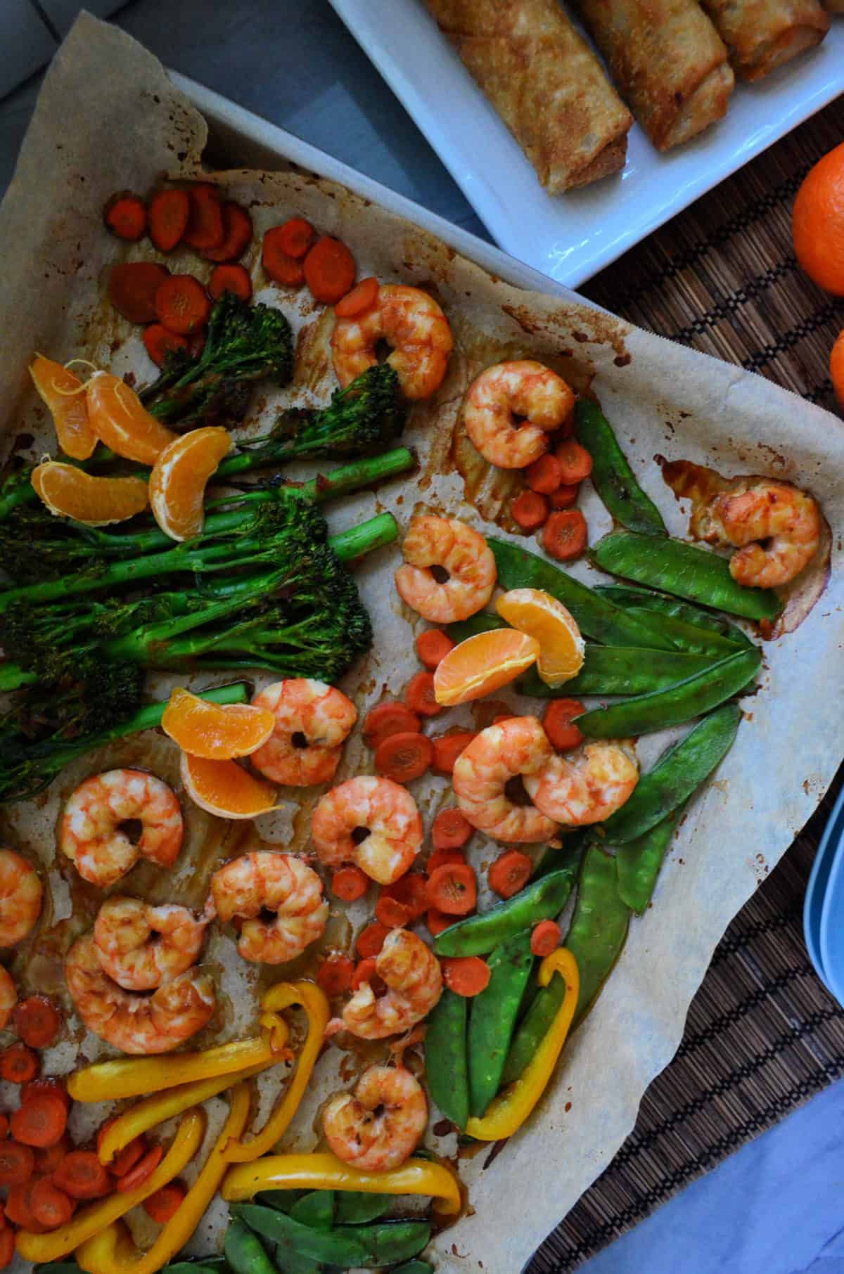 closeup peas, broccolini, mandrine oranges, shrimp, bell pepper, and carrots on paper on baking sheet.