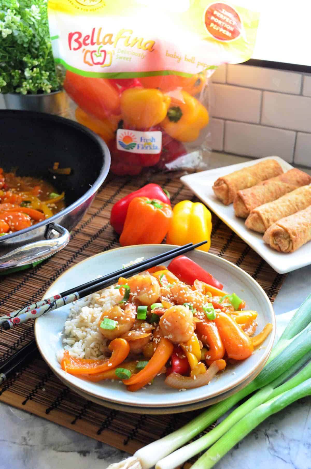 Plated Shrimp and Sweet Pepper Stir-Fry with rice in front of wok, peppers, and egg roll platter.