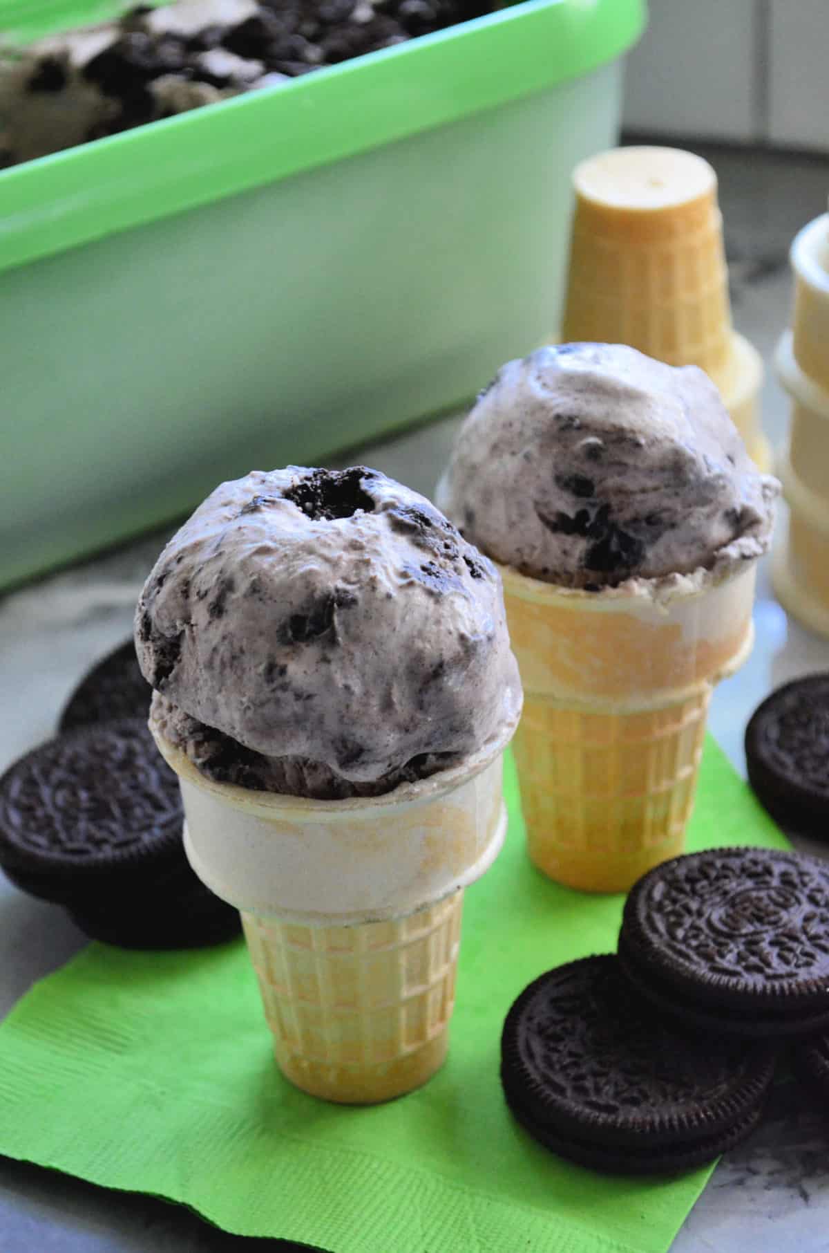 Two cake cones filled with Cookies and Cream Ice Cream with cones and oreo cookies in background.