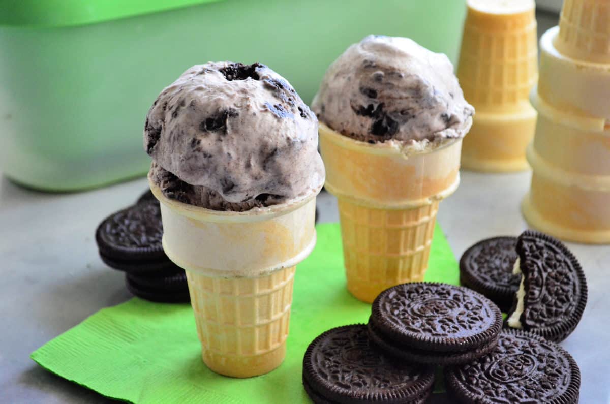 Two ice cream cones topped with oreo ice cream on a green napkin with OREO cookies next to it.