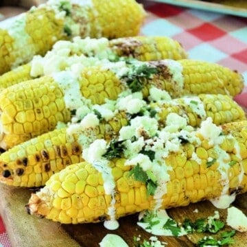side view Grilled Mexican Street Corn resting on wooden board on picnic tablecloth.