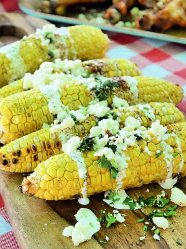 side view Grilled Mexican Street Corn resting on wooden board on picnic tablecloth.