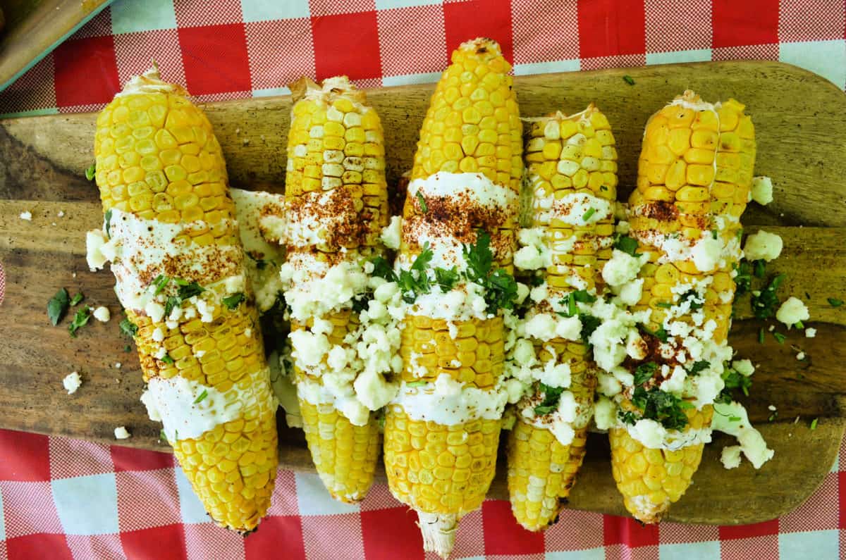 horizontal view of corn cobs on a board drizzled with crema, cheese crumbles, and herbs.