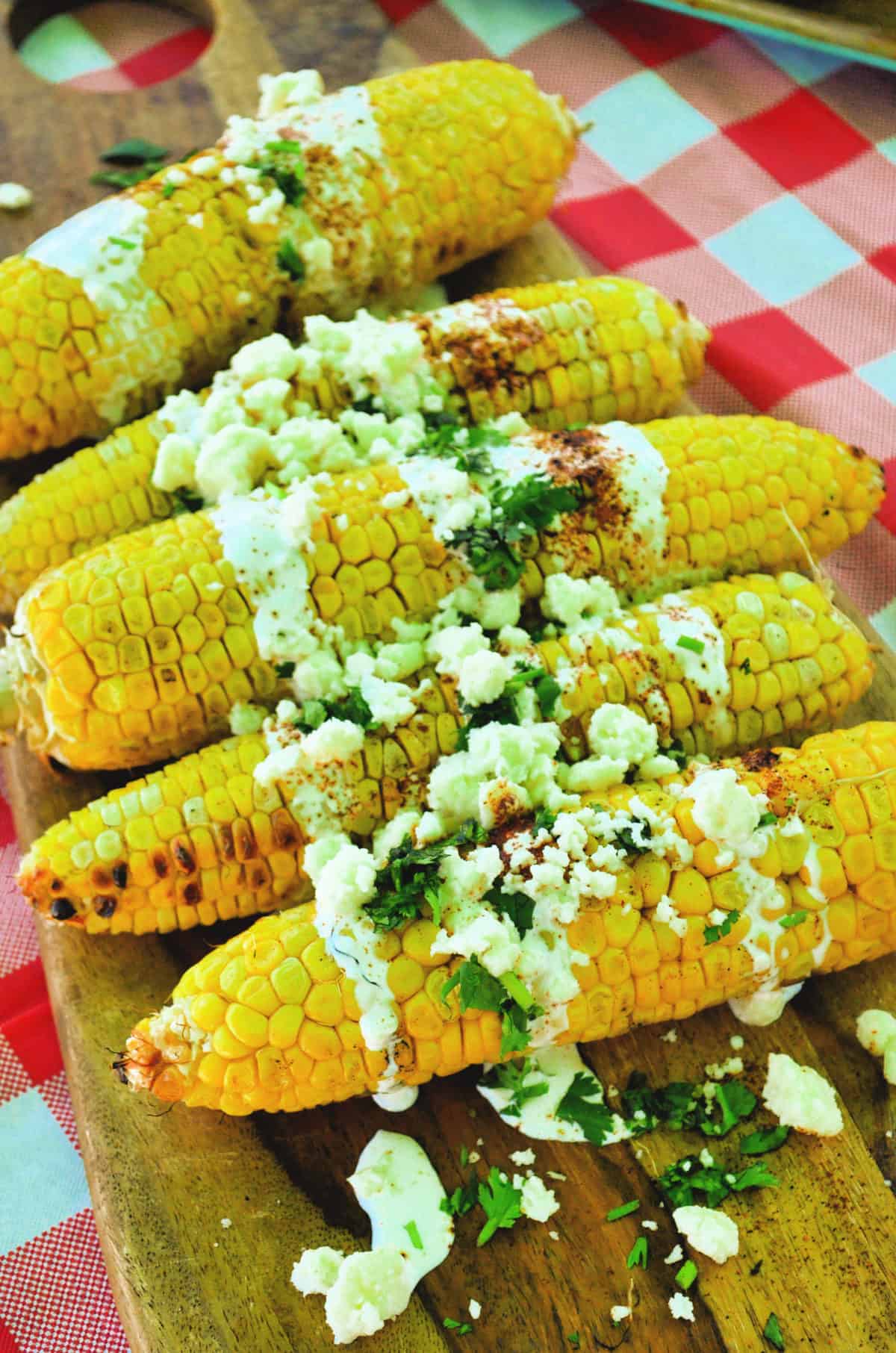 corn cobs on a board drizzled with crema, cheese crumbles, red powder, and herbs.