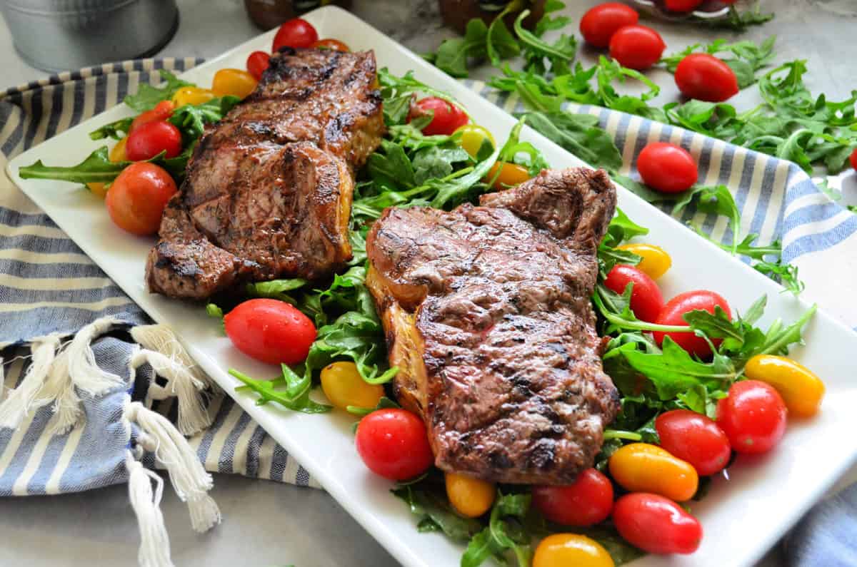 2 cooked steaks over bed of arugula salad with grape tomatoes on rectangular platter on tablelcoth.