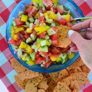 Bowl of diced watermelon, mango, onion, cilantro with hand dipping a chip into it.