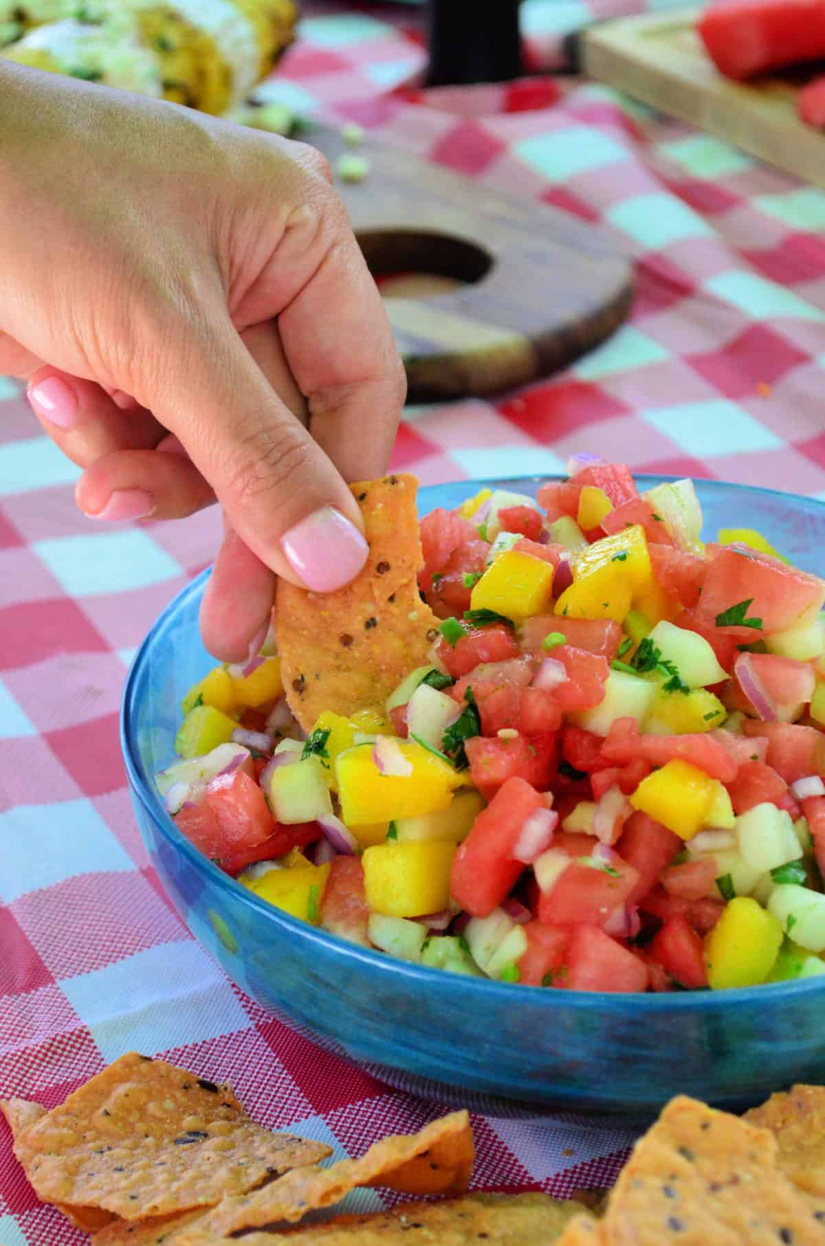 hand dipping chip into chopped watermelon with mango, red onion, and herbs most visible.