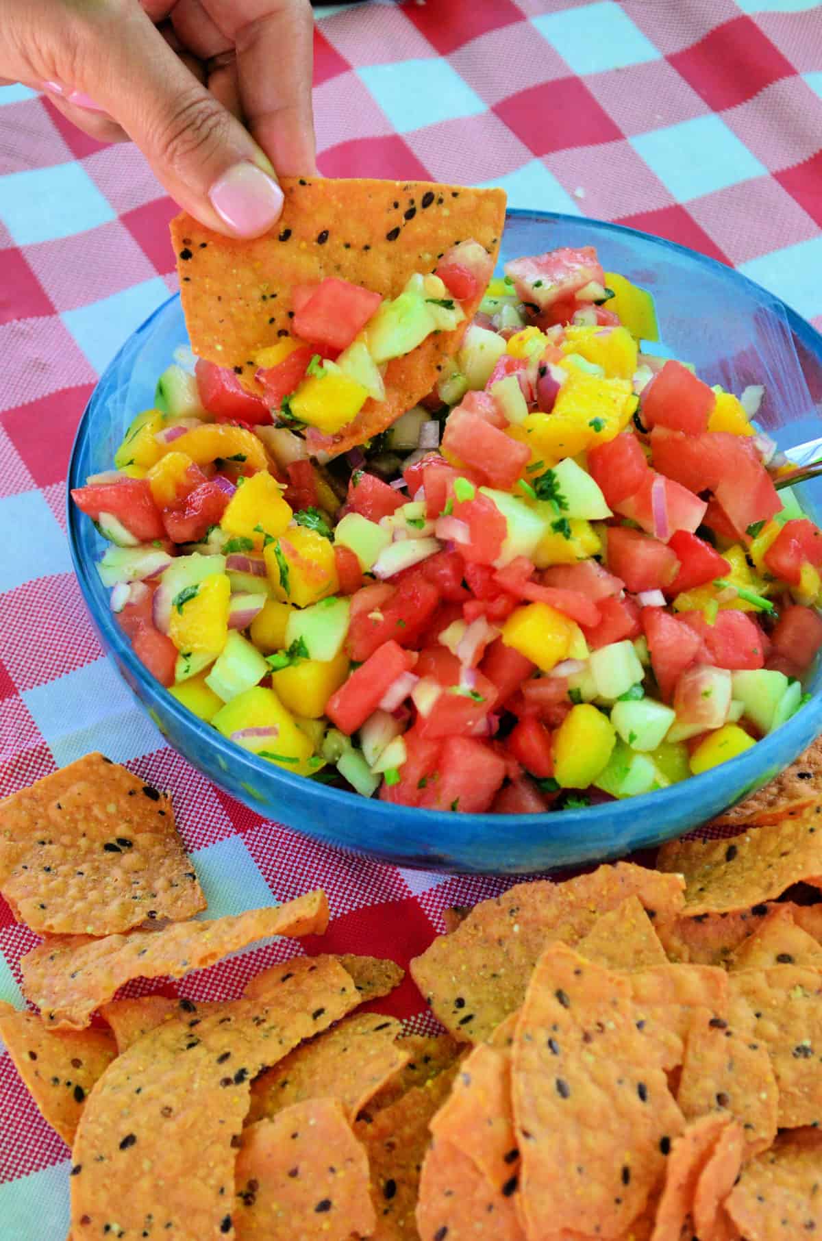 Hand dipping chip into watermelon salsa with red onions, mangos, watermelon and green herbs most visible.